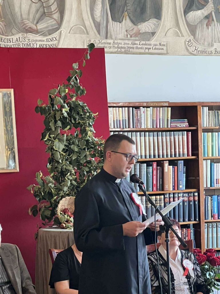 Ksiądz Damian Okroj podczas Narodowego Czytania. Stoi przed publicznością i recytuje tekst pieśni Jana Kochanowkiego.