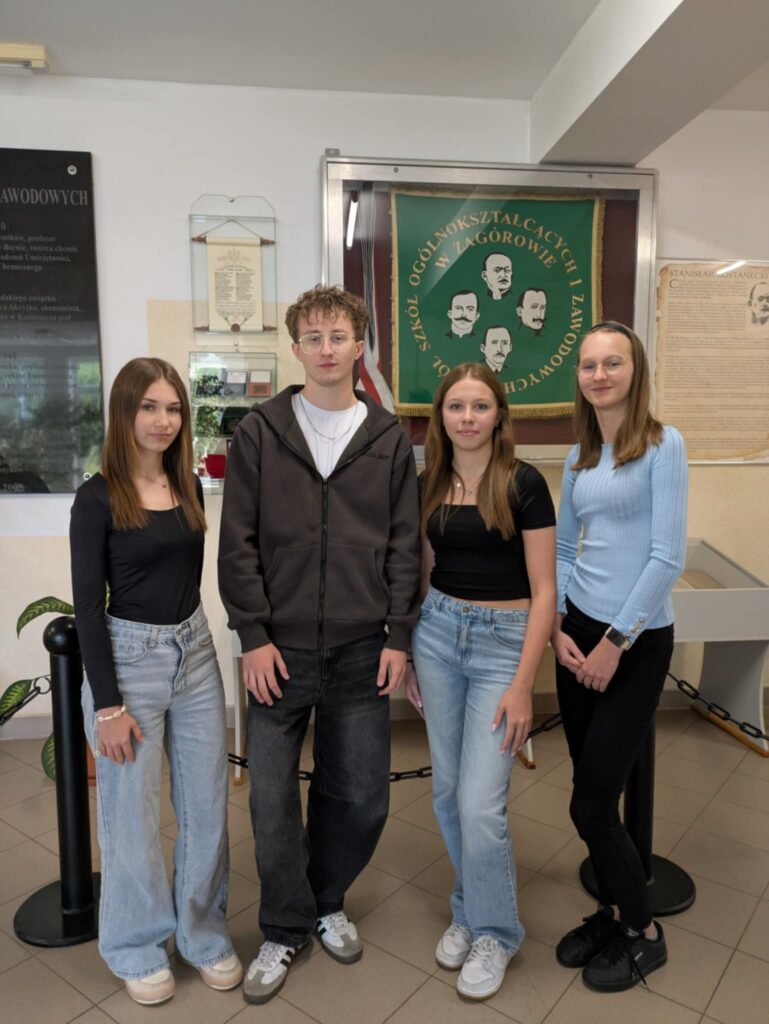 Zuzanna Zielińska, Mateusz Bryk, Natalia Matuszak i Martyna Zawal stoją razem, pozując do zdjęcia na tle szkolnego sztandaru.