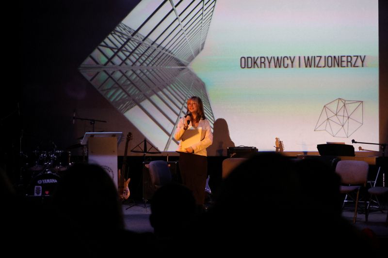 Na zdjęciu widzimy uczennicę, Alicję Waszak, stojącą na scenie podczas programu artystycznego. Scena jest oświetlona reflektorami, a w tle widać dekoracje nawiązujące do tematyki wydarzenia.