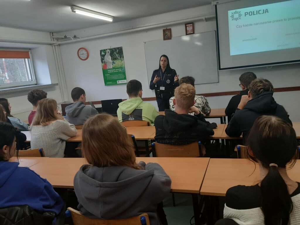Policjantka rozmawia z uczniami siedzącymi w ławkach szkolnych, w tle wyświetlana jest prezentacja.