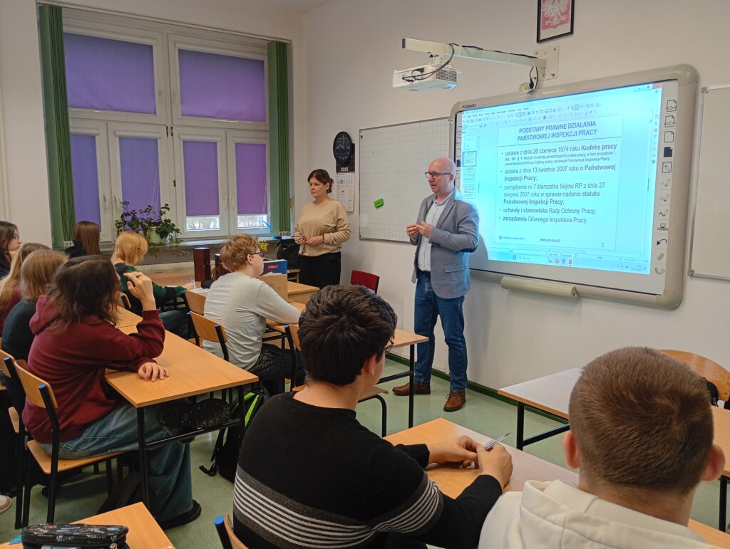 Dwoje pracowników Inspekcji Pracy prowadzi wykład stojąc przed grupą uczniów siedzących w ławkach.
