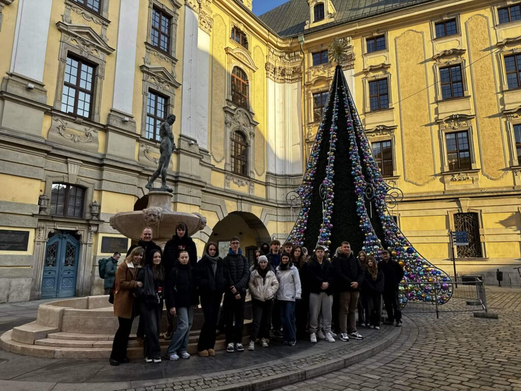 Grupa osób stoi przy zabytkowej fontannie i ozdobionej kolorowymi bombkami konstrukcji choinki na dziedzińcu historycznego budynku.