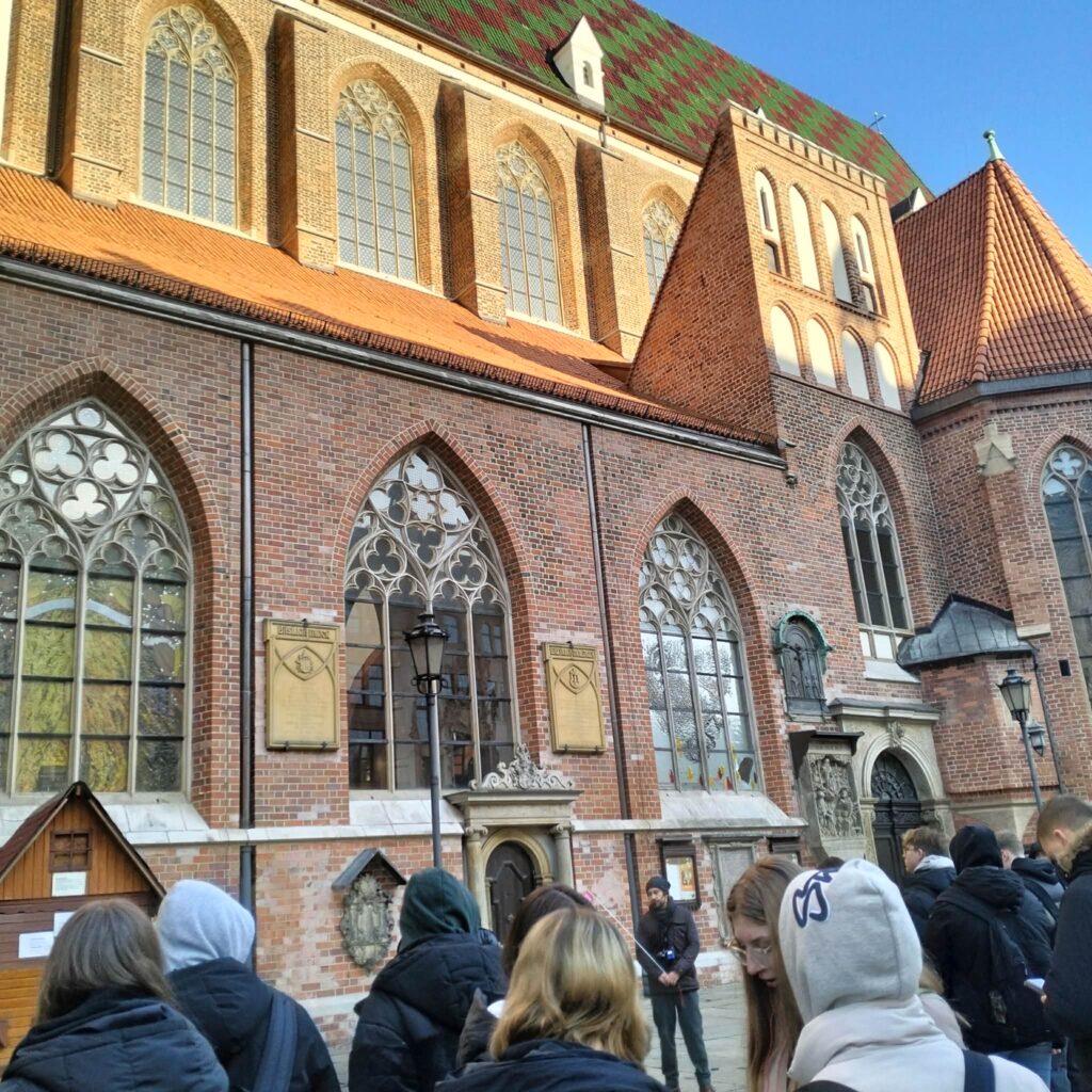 Grupa osób stoi przed zabytkowym kościołem z wielkimi gotyckimi oknami i czerwono-zielonym dachem w słoneczny dzień.