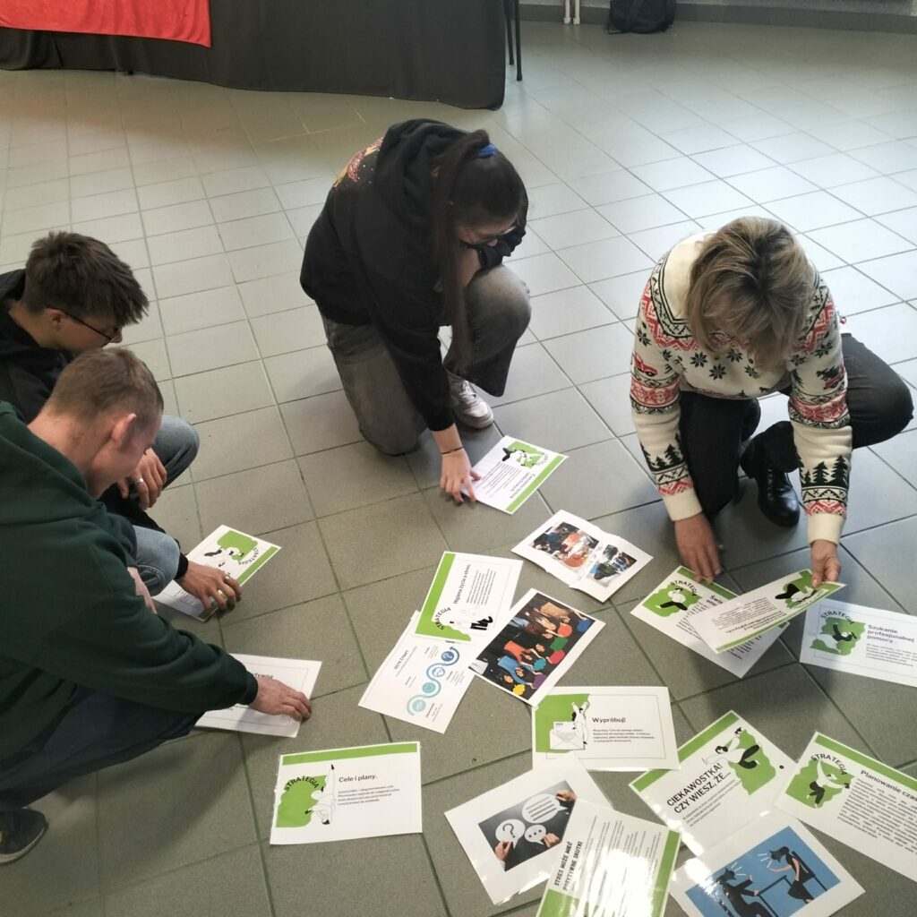 Cztery osoby klęczą na podłodze i układają karty z informacjami i grafikami dotyczącymi strategii i planowania.