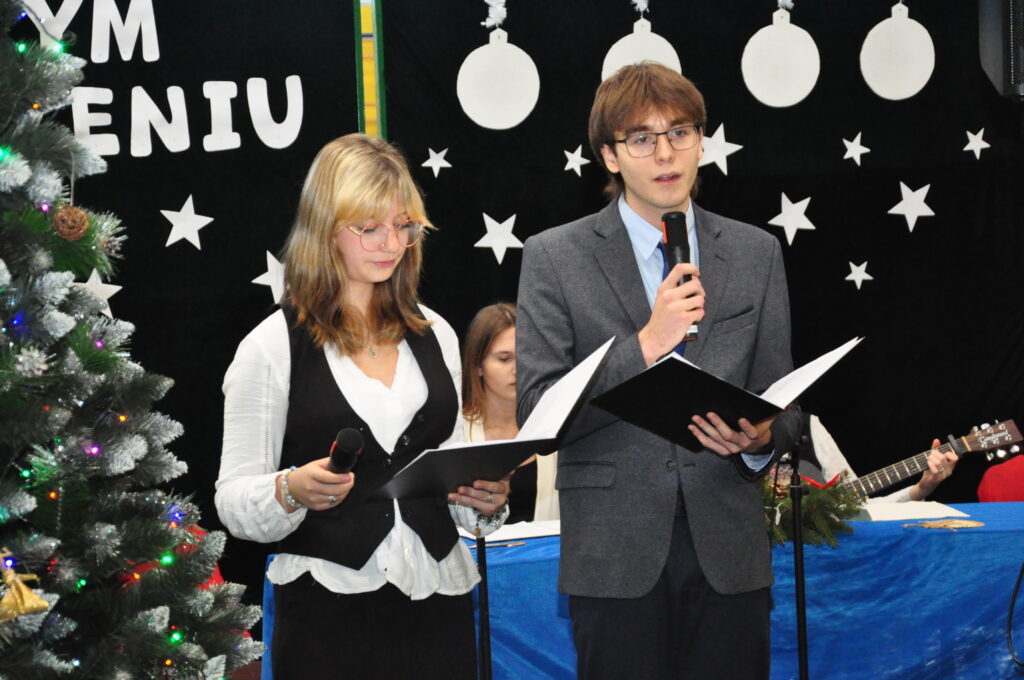 Dwóch młodych ludzi ubranych elegancko czyta z teczek, trzymając mikrofony podczas świątecznego wydarzenia.