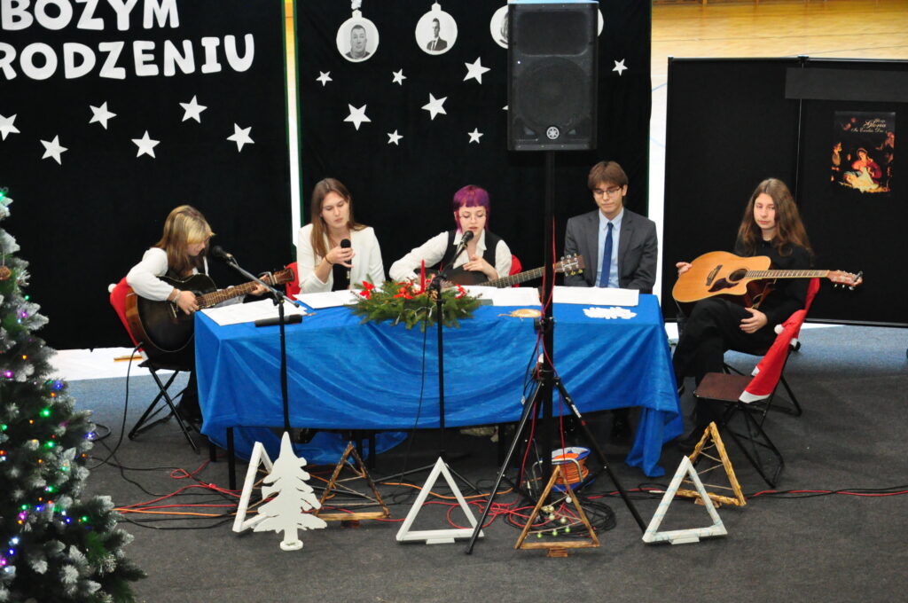 Młodzi ludzie siedzą przy stole ozdobionym świątecznym stroikiem, grają na gitarach i śpiewają podczas bożonarodzeniowego wydarzenia.