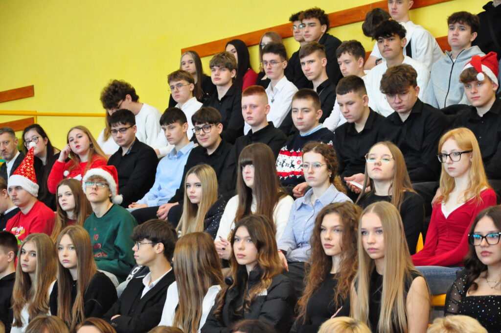 Grupa młodych osób w różnych ubraniach, niektórzy w świątecznych czapkach, siedzą w widowni na żółtych schodach.