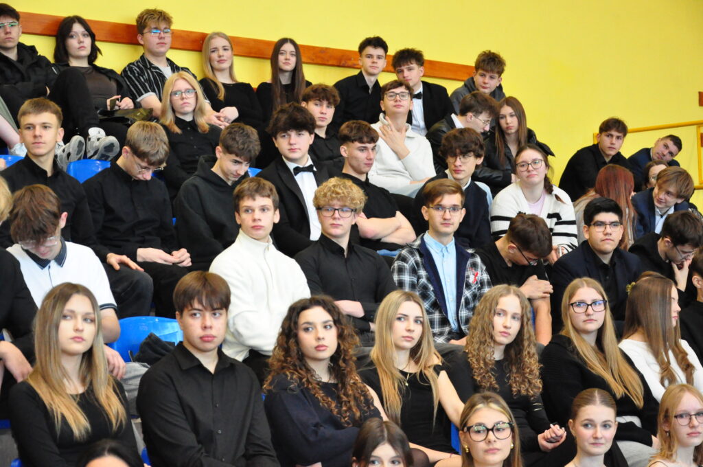 Grupa młodych osób siedzi na trybunach w sali z żółtymi ścianami, większość ubrana na czarno, niektórzy mają okulary.