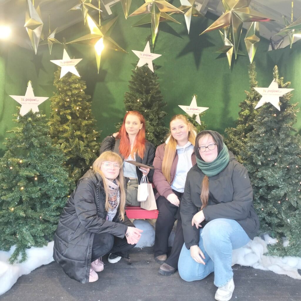 Cztery młode kobiety pozują w świątecznej dekoracji z zielonymi choinkami i dużymi złotymi gwiazdami.