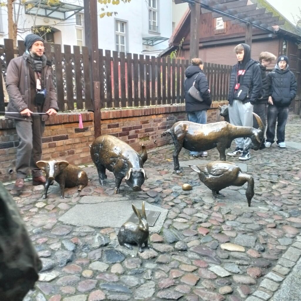 Figurki zwierząt gospodarskich wykonane z brązu ustawione na brukowanym chodniku, obok nich stoją ludzie.