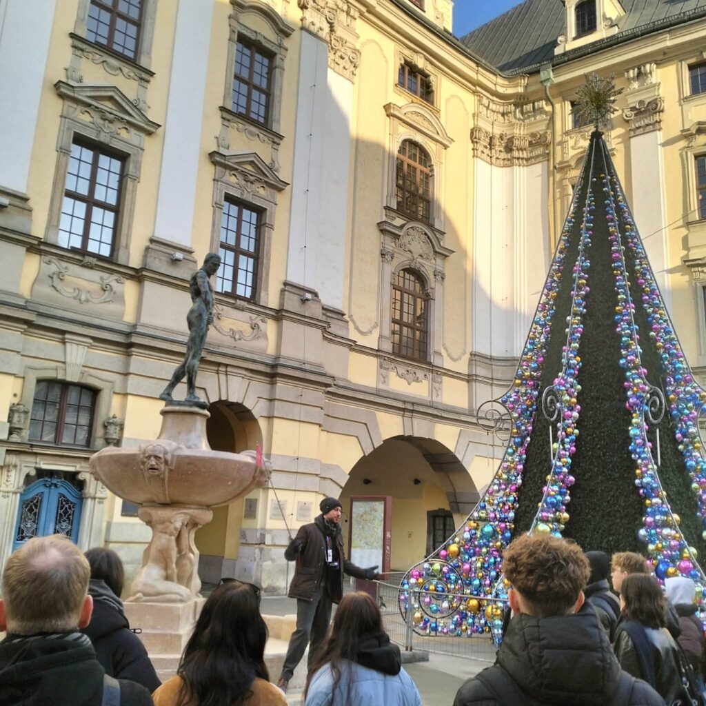 Grupa ludzi słucha przewodnika stojącego obok fontanny i dużej choinki udekorowanej kolorowymi bombkami na tle renesansowego budynku.