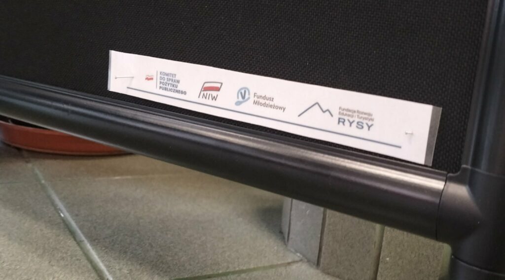 Logotypy i nazwy organizacji: Komitet do Spraw Pożytku Publicznego, NIW, Fundusz Młodzieżowy oraz Fundacja Rozwoju Edukacji i Turystyki RYSY na fragmentu tabliczki mocowanej do czarnej powierzchni.