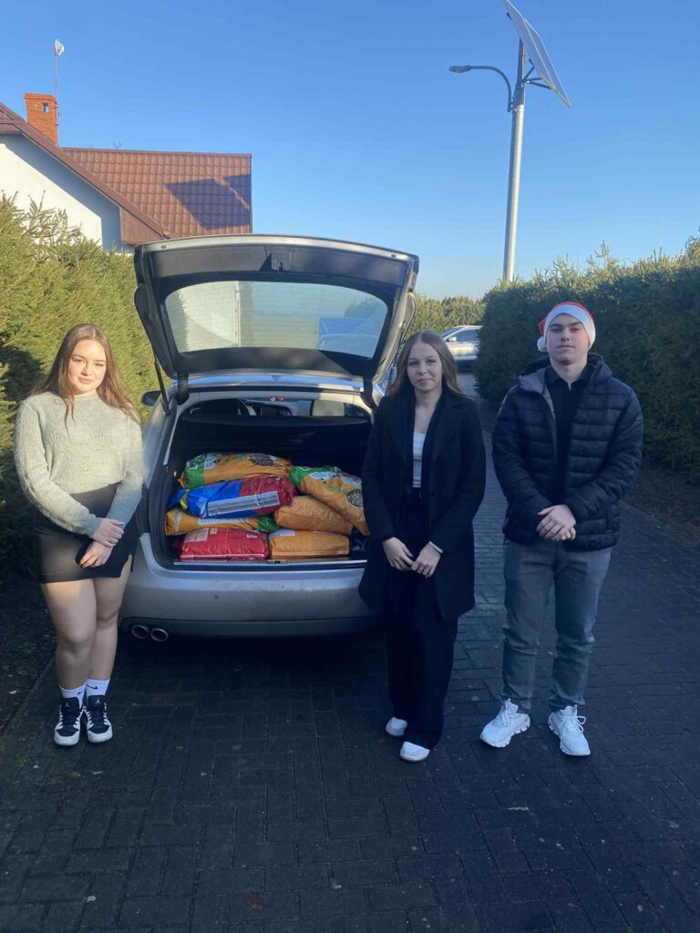 Trzy młode osoby stoją przed otwartym bagażnikiem samochodu wypełnionym dużymi workami karmy.
