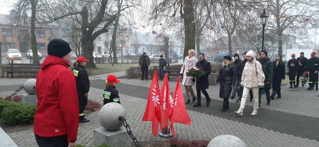 Grupa osób idzie w uroczystym pochodzie, niesąc wiązankę kwiatów przed pomnikiem z czerwonymi flagami, a strażacy w mundurach stoją na baczność.