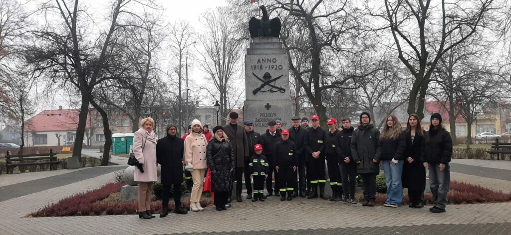 Grupa osób różnych pokoleń, w tym strażacy w mundurach, stoi razem przy pomniku upamiętniającym poległych w latach 1918-1920.