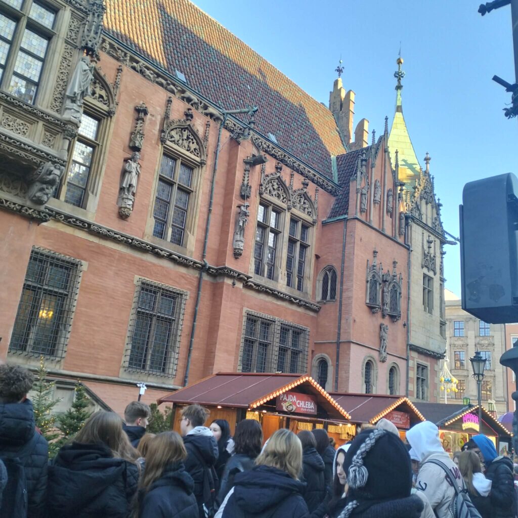 Tłum ludzi stoi przed stoiskami z jedzeniem na tle zabytkowego, gotyckiego budynku z czerwonej cegły ozdobionego rzeźbami i dekoracyjnym dachem.