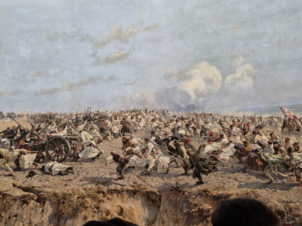 Scena historycznej bitwy z żołnierzami w białych strojach atakującymi okopy i działo na polu walki.