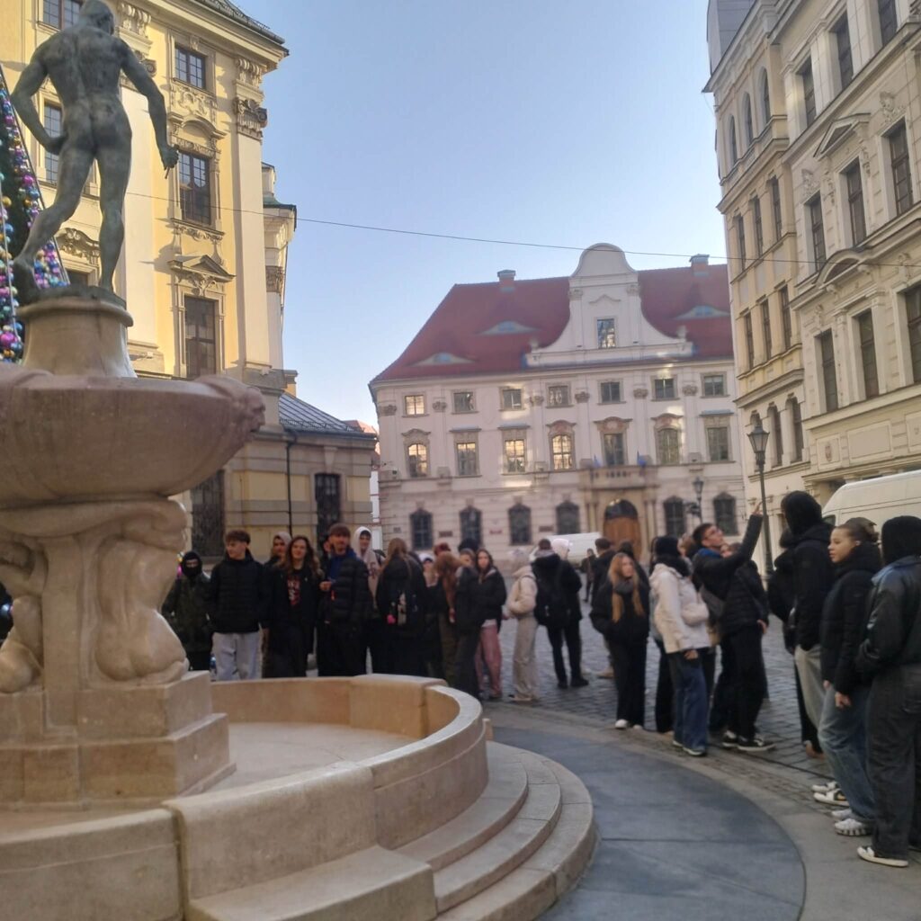 Grupa ludzi stoi w kolejce obok kamiennej fontanny z rzeźbą nagiego mężczyzny, w tle zabytkowe budynki z czerwonym dachem.