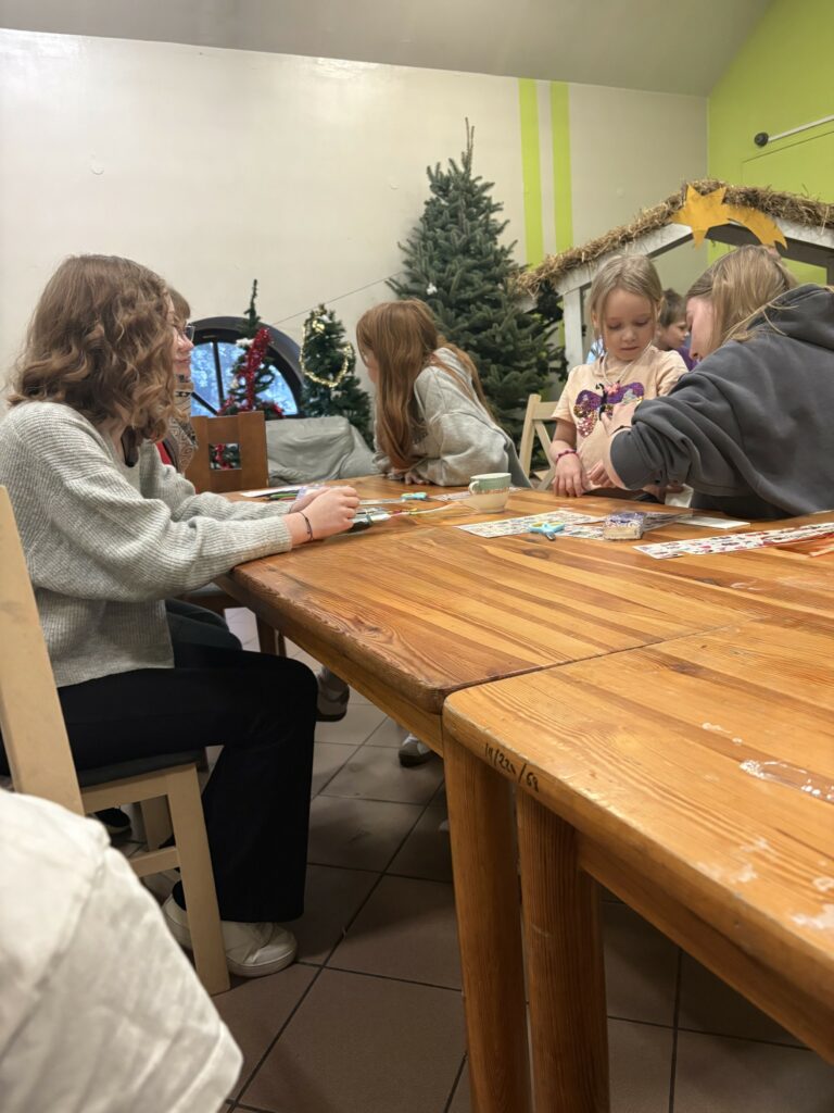 Grupa dzieci i młodzieży siedzi przy drewnianym stole, wykonując prace plastyczne w świątecznie udekorowanym pomieszczeniu.