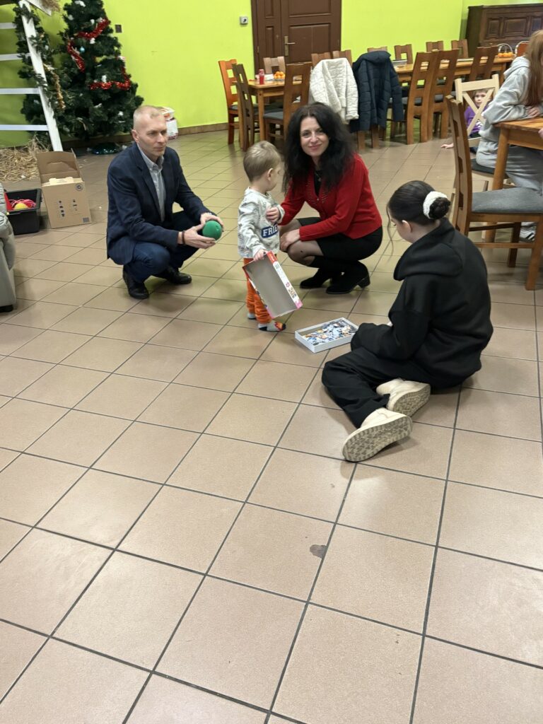 Dorośli i dzieci bawią się razem na podłodze przy świątecznie udekorowanym pokoju z choinką i stołami.