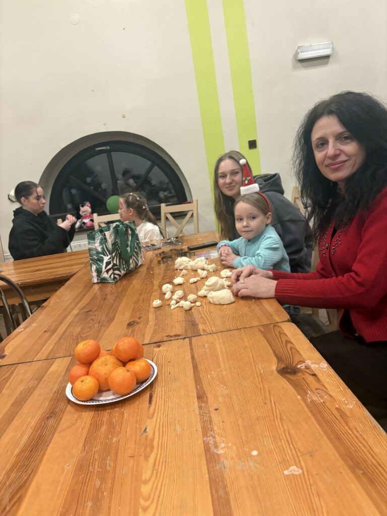 Grupa osób, w tym dzieci, siedzi przy stole i formuje masę plastyczną, na stole leży talerz mandarynek.