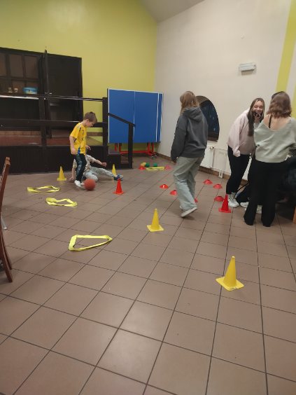 Dzieci uczestniczą w zabawie z piłką i pachołkami na sali z żółtymi i czerwonymi przeszkodami ułożonymi na podłodze.