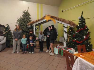 Grupa dzieci i młodzieży stoi obok drewnianej szopki bożonarodzeniowej z gwiazdą na dachu, obok choinek i prezentów.