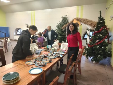 Ludzie przygotowują świąteczne spotkanie przy stole z filiżankami i ciastem, obok stoi udekorowana choinka i szopka bożonarodzeniowa.