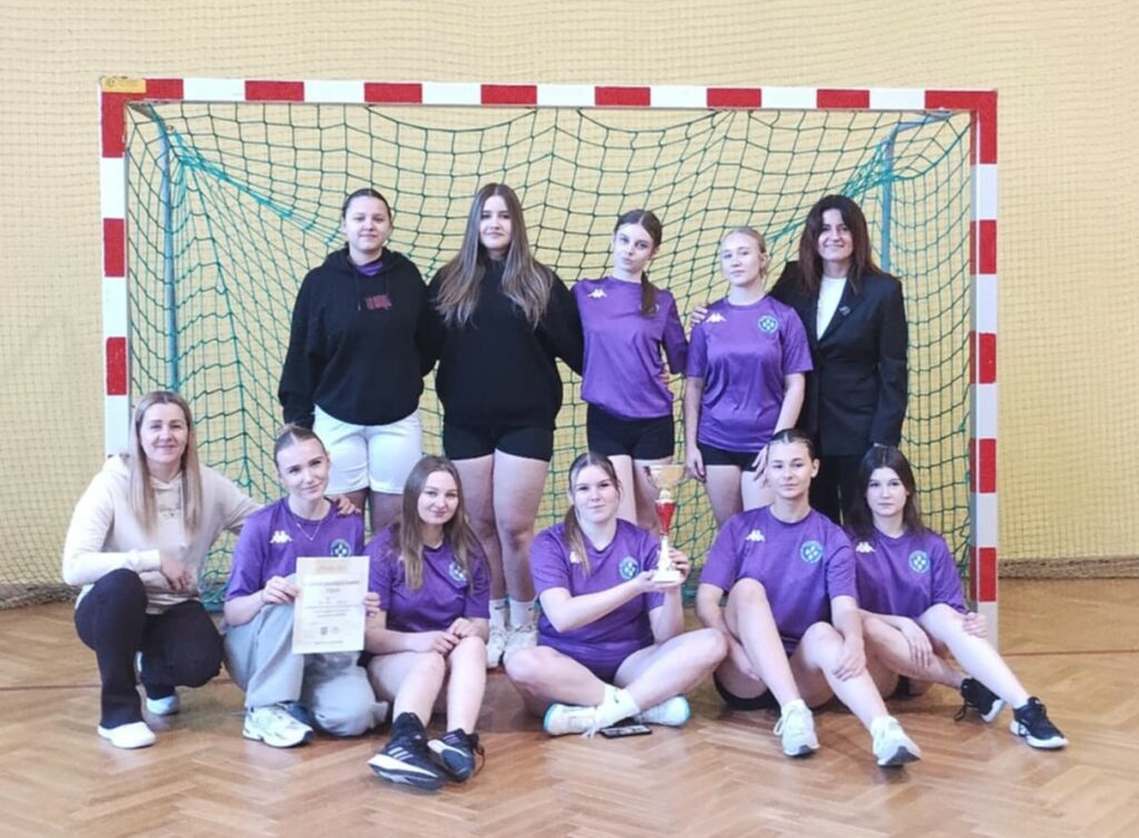 Dziewczęca drużyna z pucharem i dyplomem, pozująca na tle bramki w hali sportowej.