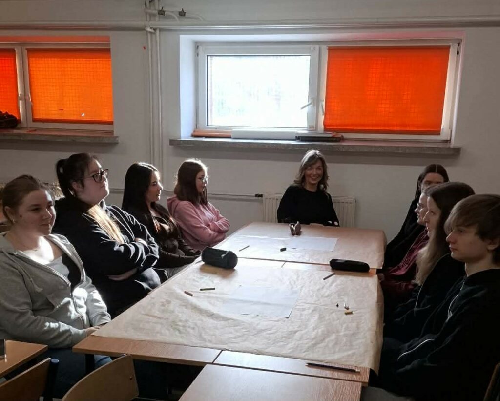 Grupa młodzieży siedzi przy dwóch stolikach ustawionych razem w klasie z jasnymi oknami zasłoniętymi pomarańczowymi roletami.