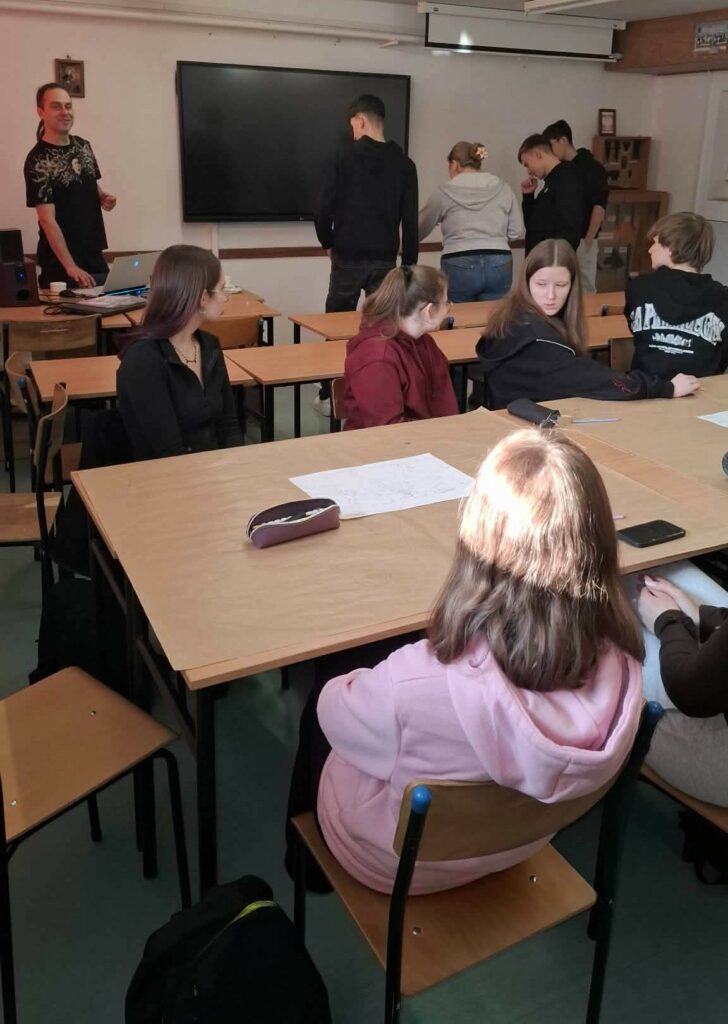 Grupa młodych osób siedzi i stoi przy stołach w sali lekcyjnej, a nauczyciel uśmiecha się stojąc przy biurku z laptopem.