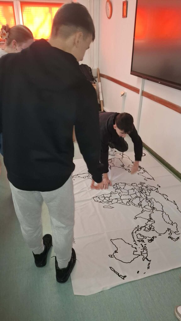 Młodzi ludzie z zaangażowaniem wskazują różne państwa na dużej, rozłożonej w pomieszczeniu mapie konturowej świata.