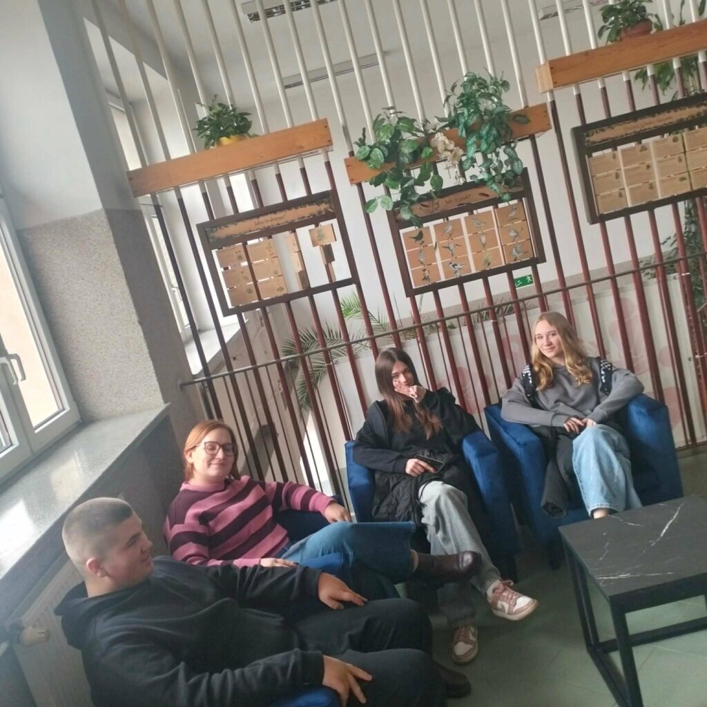 Cztery osoby siedzą na niebieskich fotelach w przyjaznym wnętrzu z roślinami i tablicami edukacyjnymi na ścianie.