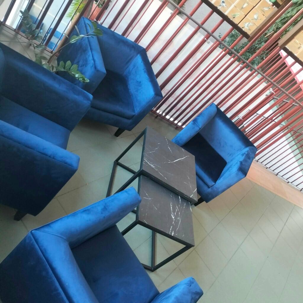 Zestaw czterech niebieskich foteli wokół dwóch kwadratowych stolików z czarnymi blatami o marmurowym wzorze, ustawionych na jasnej podłodze obok drewnianych barier.