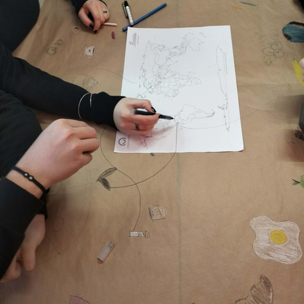 Dwie osoby rysują na brązowym papierze rozłożonym na stole, z mapą świata i różnymi przyborami do rysowania.
