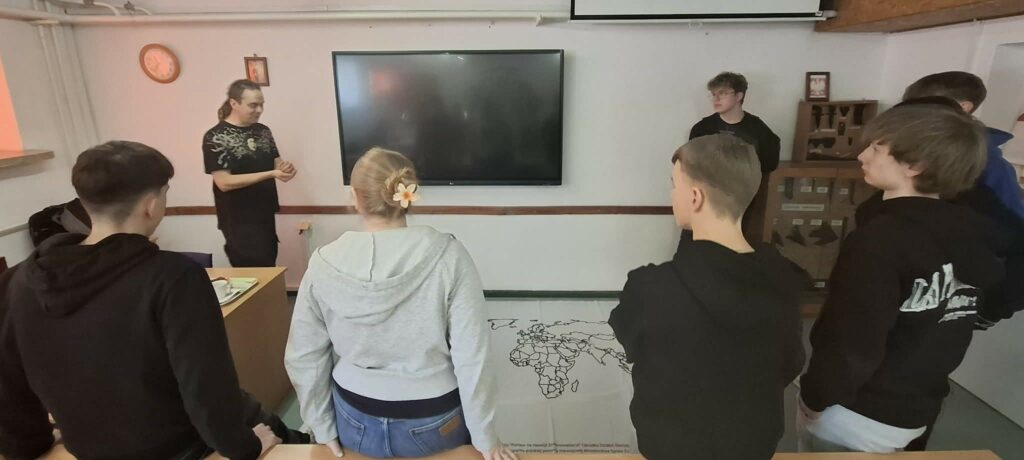 Grupa młodzieży uczestniczy w lekcji lub prezentacji w klasie, podczas której nauczyciel stoi obok dużego ekranu i mapy świata rozłożonej na podłodze.
