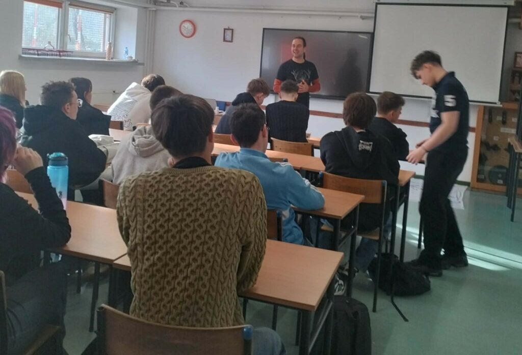 Grupa młodzieży siedzi w ławkach szkolnych, słuchając nauczyciela prowadzącego lekcję przy tablicy multimedialnej.