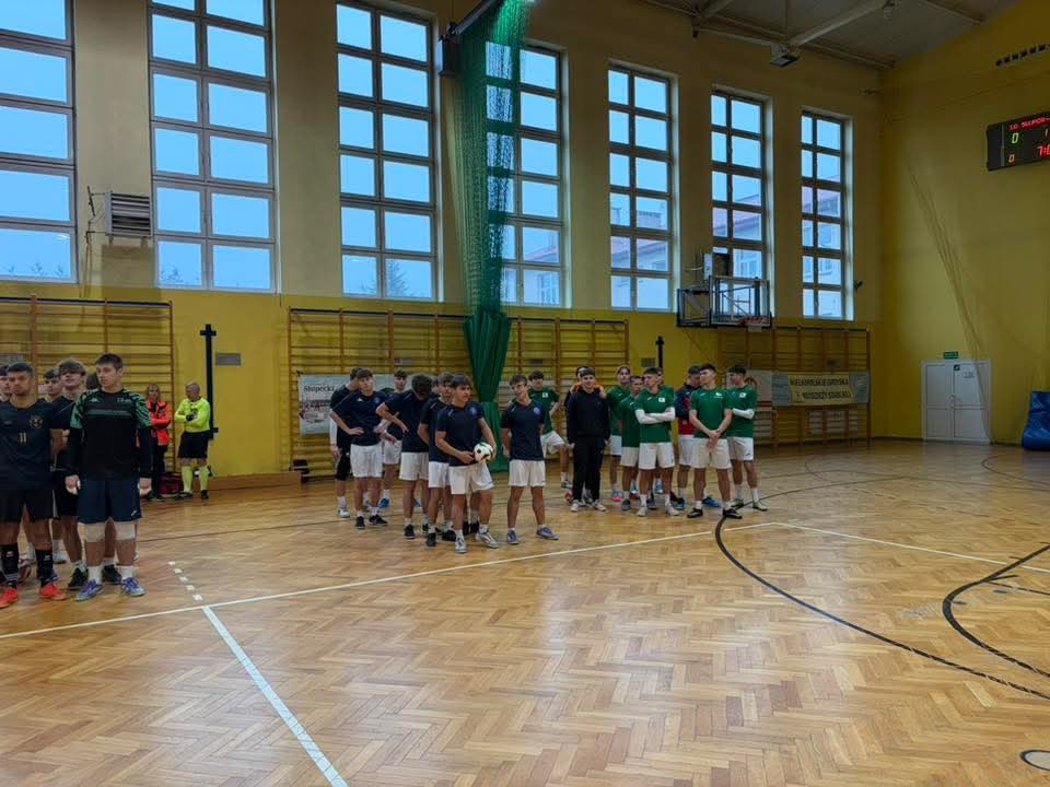 Drużyny piłkarskie w hali sportowej ustawione w szeregi przed rozpoczęciem meczu, jedna w strojach czarnych, druga w zielonych.