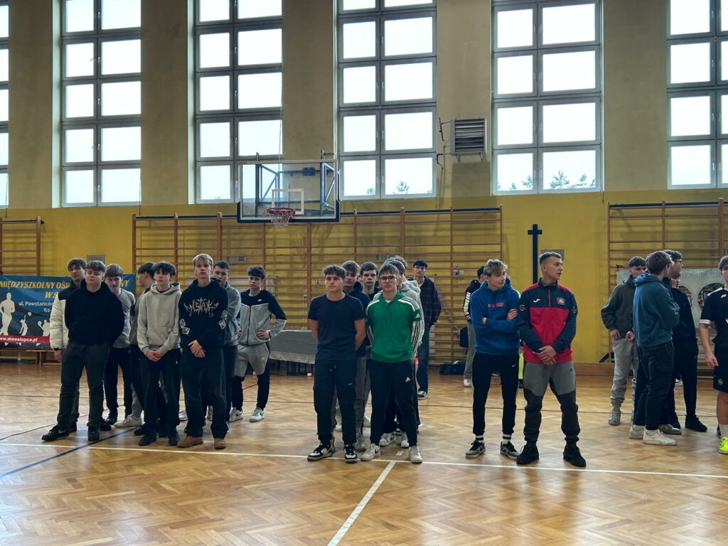 Grupa młodych mężczyzn stoi w hali sportowej, ustawionych w kilku rzędach przed koszem do koszykówki.