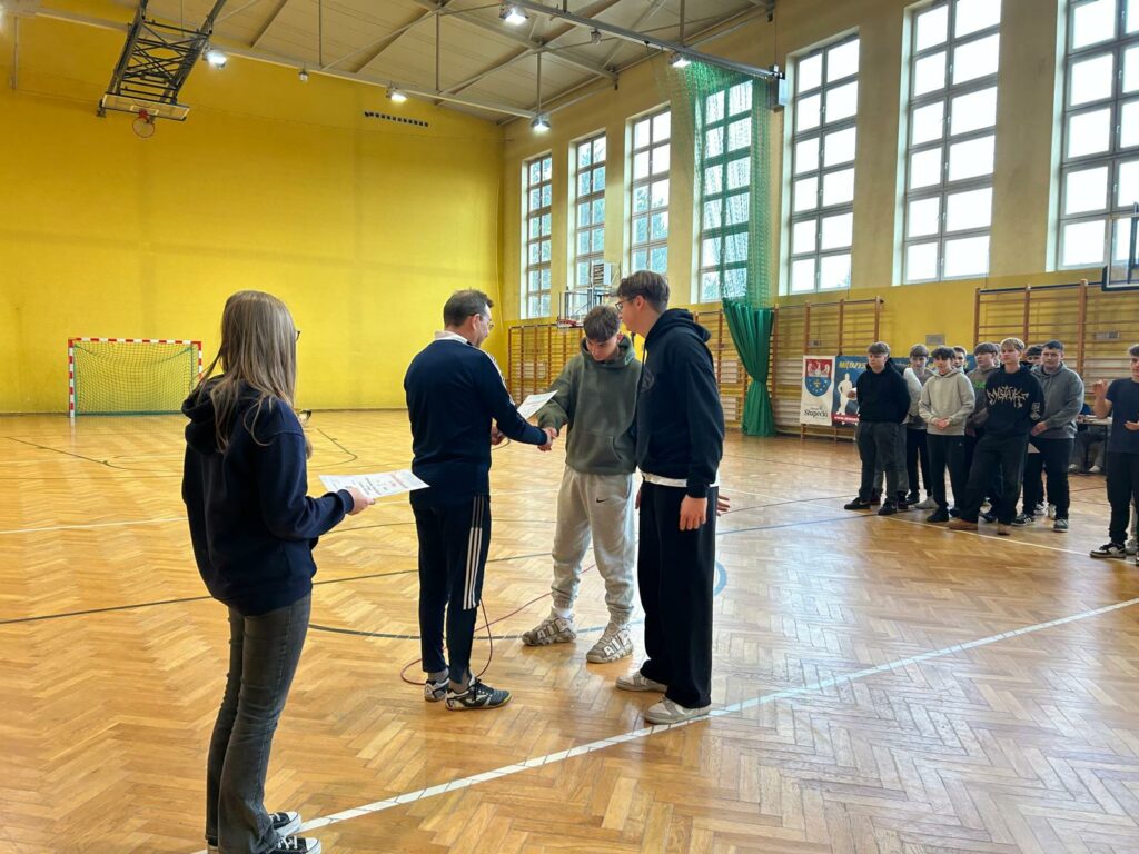 Grupa młodzieży stoi w hali sportowej, gdzie jedna z osób wręcza innym dyplomy i gratuluje, podczas gdy reszta grupy oczekuje w kolejce.