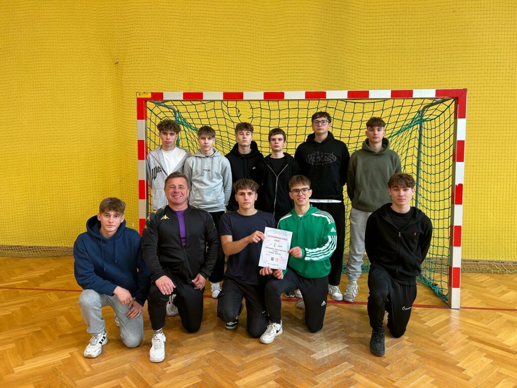 Dziesięciu młodych mężczyzn i dorosły trener pozują na tle bramki w hali sportowej, trzech z nich trzyma dyplom.