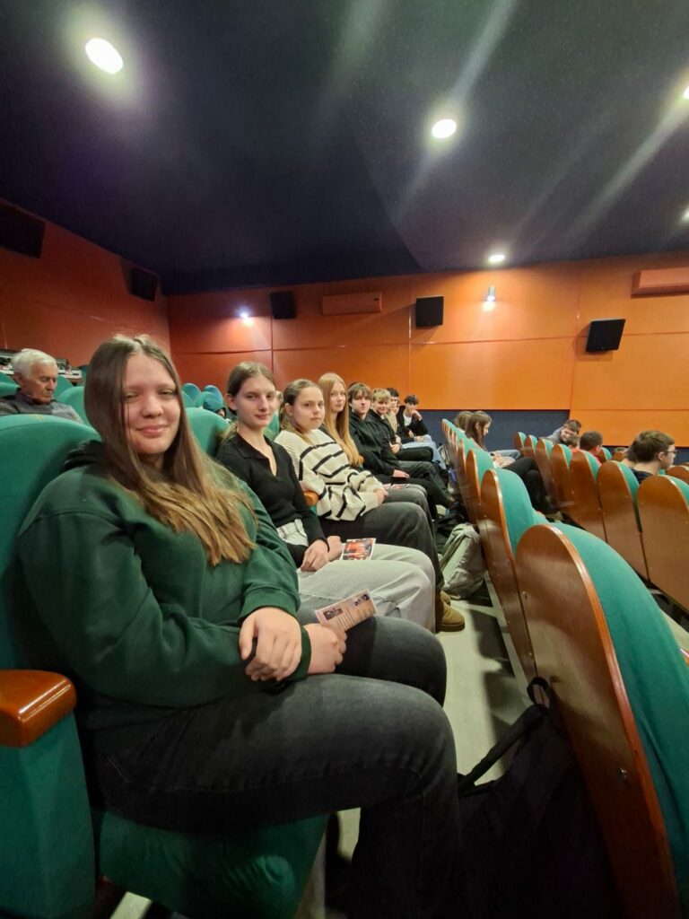 Grupa młodych osób siedzi obok siebie w kinie lub teatrze na zielonych fotelach, trzymając w rękach programy lub broszury.