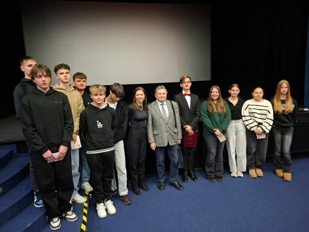 Grupa młodych osób wraz z dwoma dorosłymi stoi na scenie przed ekranem w sali kinowej lub teatralnej.