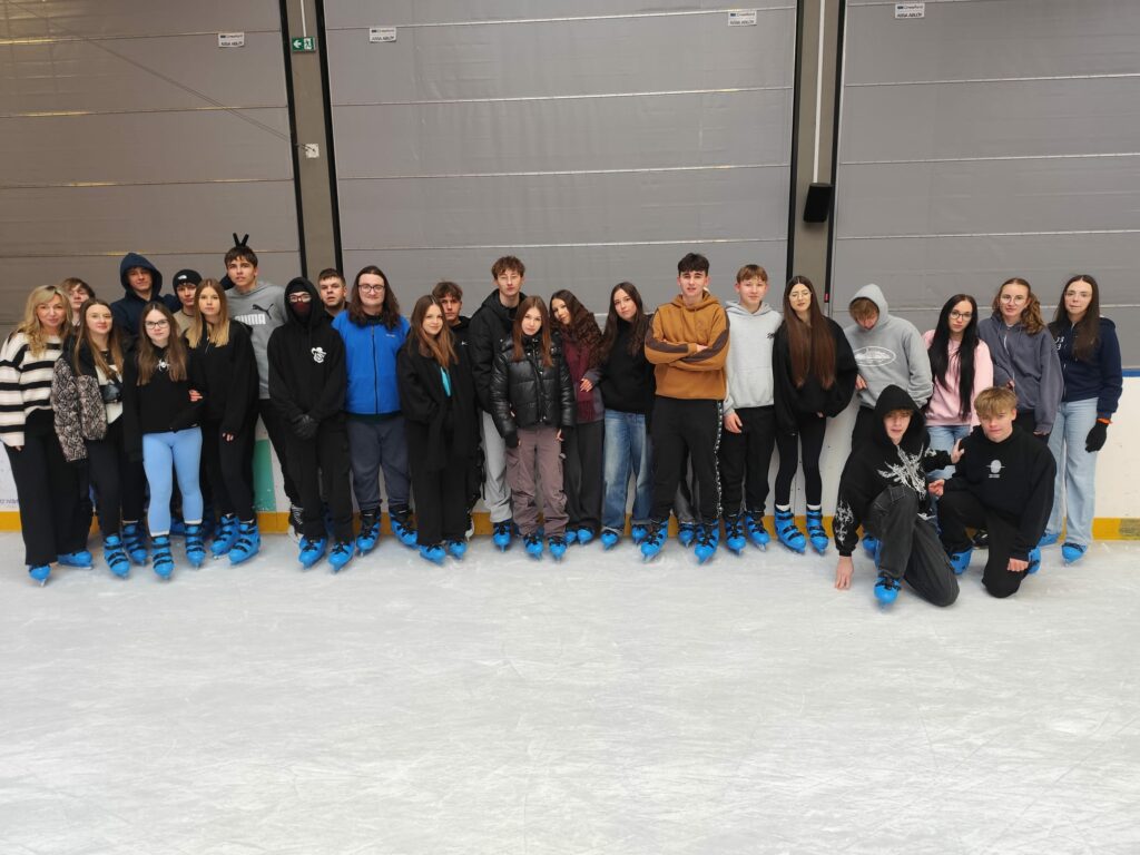 Grupa młodych osób ubranych w ciepłe ubrania i niebieskie łyżwy stoi razem na lodowisku.