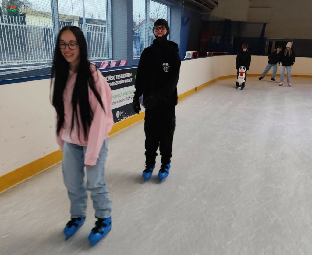 Grupa młodych osób jeździ na łyżwach na krytym lodowisku, jedna z nich korzysta z pomocy w postaci figurki pingwina.