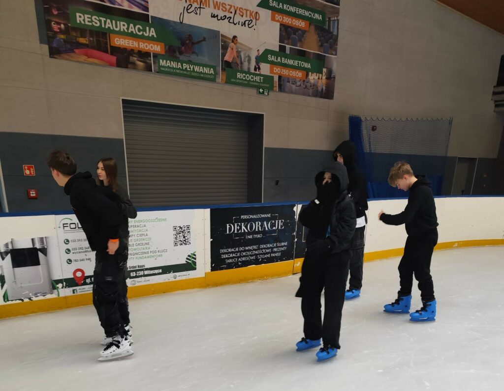 Młodzi ludzie jeżdżą na łyżwach na lodowisku, niektórzy ubrani w czarne ubrania z kapturem, a część w niebieskich łyżwach.