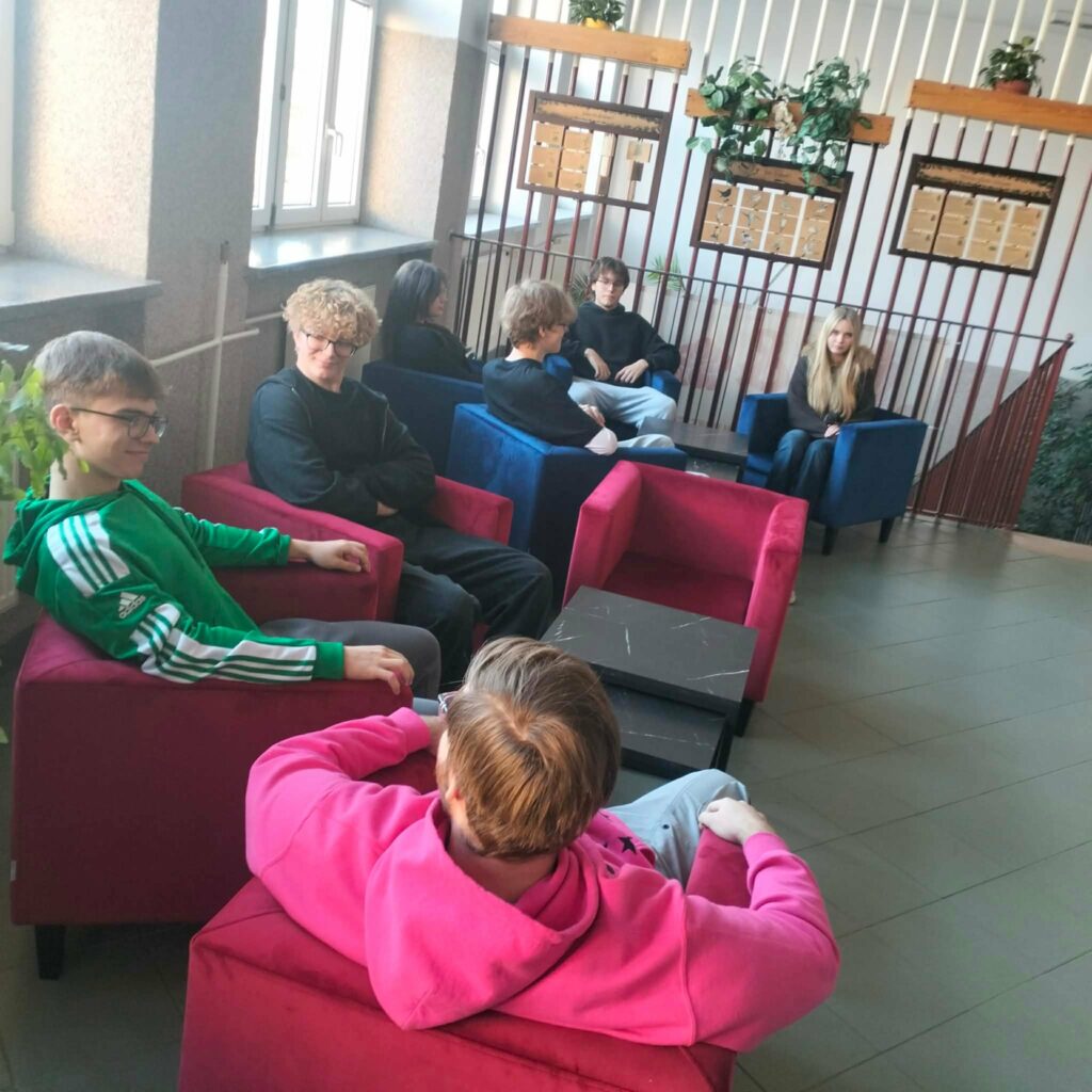 Grupa młodych osób siedzi na kolorowych fotelach w korytarzu lub poczekalni o dużych oknach i roślinach.