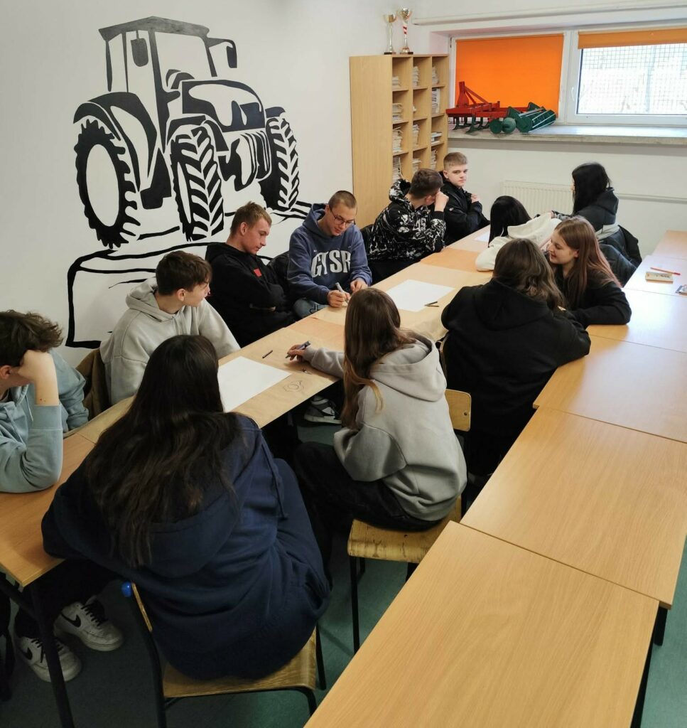 Grupa młodzieży siedzi przy stołach w klasie z rysunkami i kartkami papieru, na ścianie widoczny jest duży czarno-biały mural traktora.