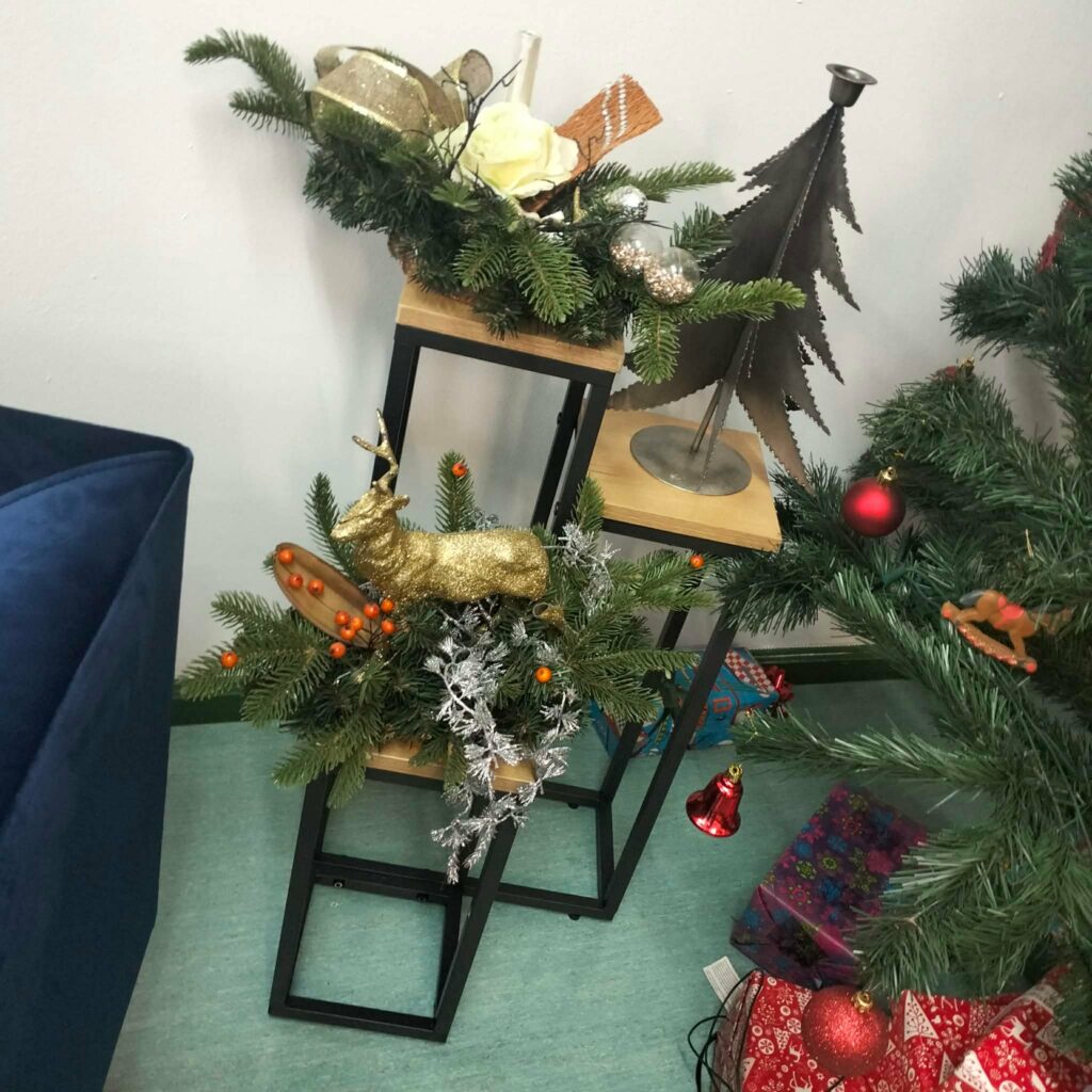 Zielone gałązki świerku ozdobione złotym reniferem, czerwonymi jagodami, srebrnymi elementami i świątecznymi dekoracjami stoją na dwupoziomowym metalowo-drewnianym stojaku obok przystrojonej choinki i prezentów.