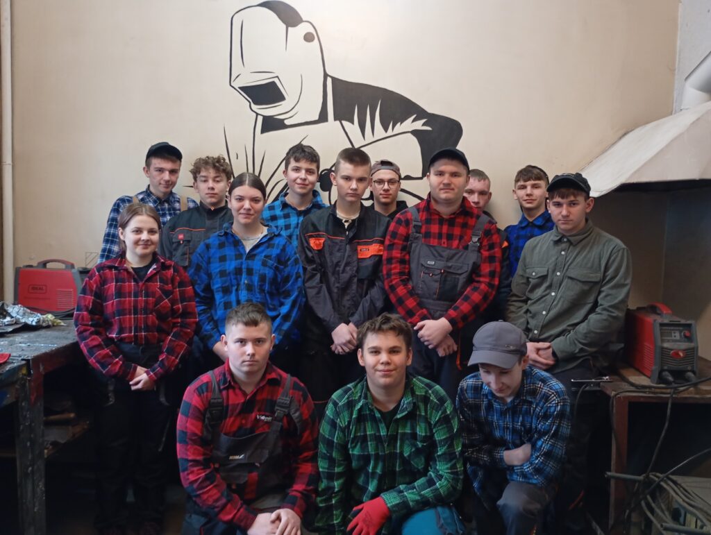 Grupa nastolatków ubrana w robocze ubrania i koszule w kratę stoi i klęczy w warsztacie z muralem spawacza w tle.